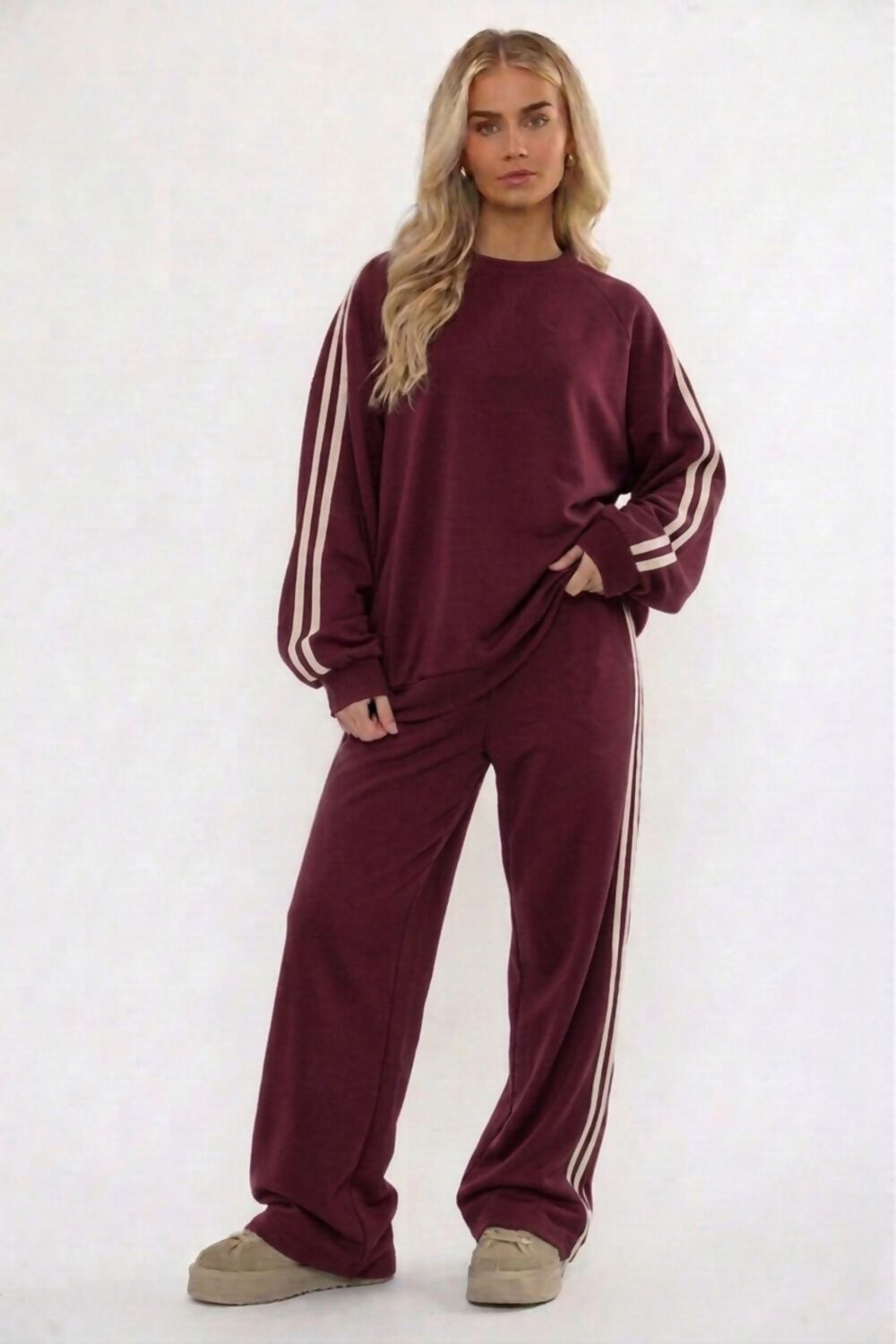 A woman modeling a matching brown lounge set consisting of a long-sleeve sweatshirt and wide-leg sweatpants, both featuring white stripes down the sleeves and legs, against a plain white studio background.