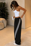 A woman modeling black wide-leg sweatpants with three white side stripes, paired with a white ribbed tank top and white sneakers, standing in a modern indoor room.