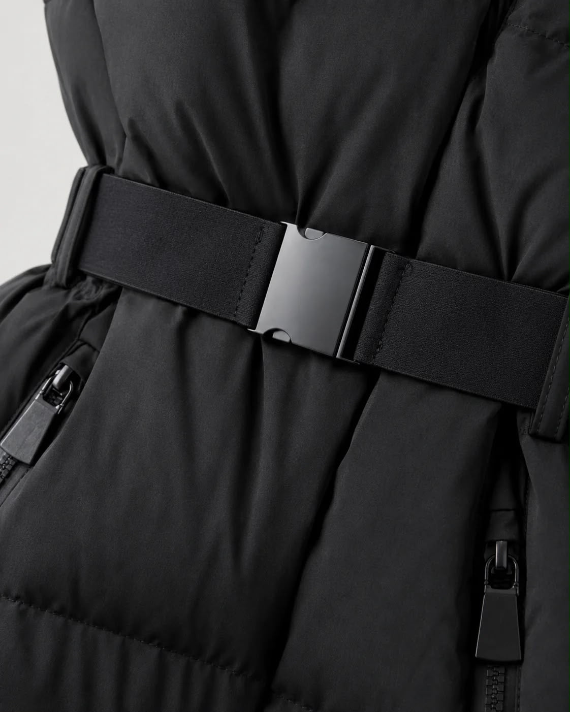 Close-up of a black puffer jacket with a wide black belt and a matte black metal buckle, showing quilted stitching and zippered pockets.