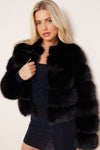 A blonde model wearing a luxurious black fur jacket over a dark glittery dress, posing against a plain off-white background.