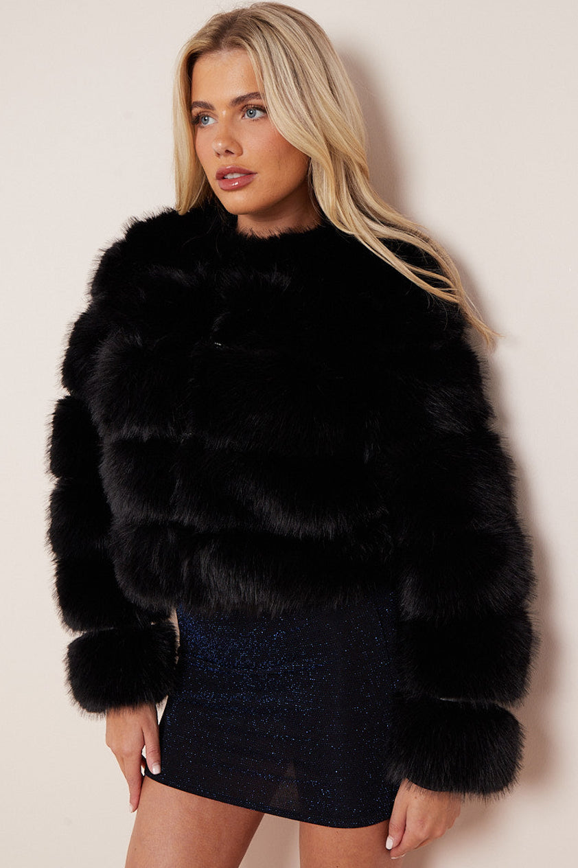 A blonde model wearing a cropped black faux fur jacket with a high collar and matching faux fur cuffs, paired with a dark blue sequined mini skirt, against a plain beige background.