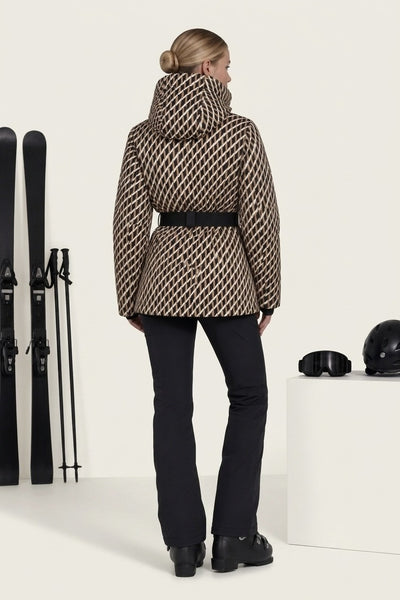 A woman stands with her back to the camera wearing a beige and black patterned ski jacket with a black belt, black ski pants, and black ski boots. Ski equipment and a white display block are visible in a clean studio setting.