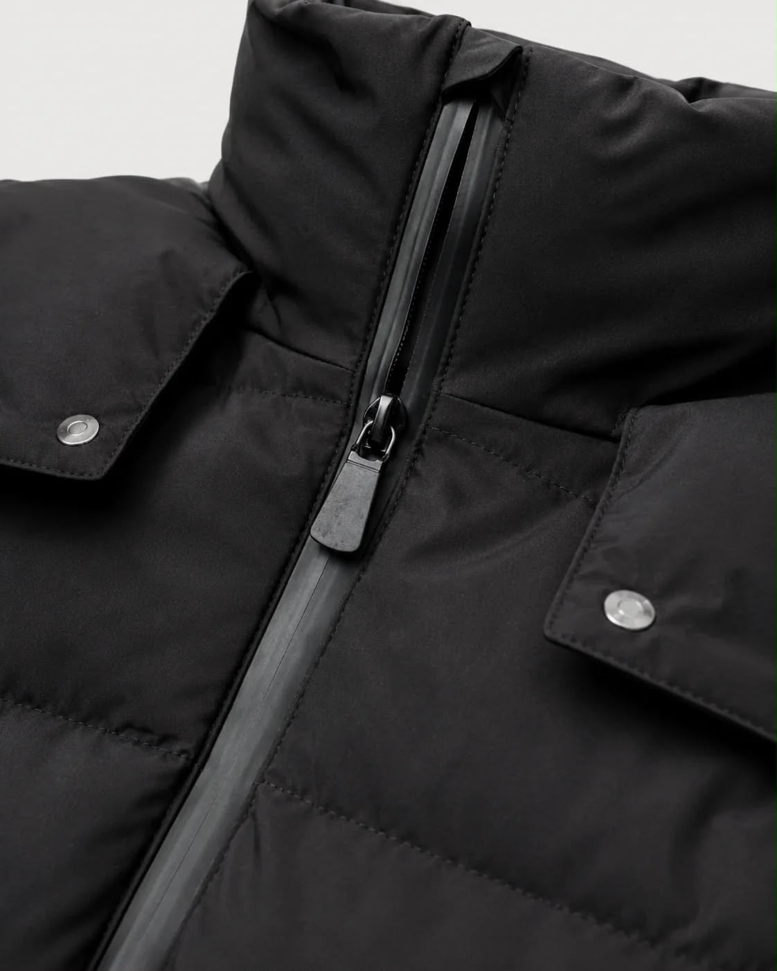 Close-up of a black puffer jacket showing the front zipper, high collar, and flap pockets with silver snap buttons.