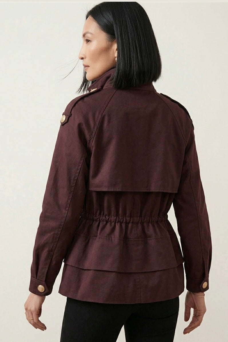 A woman shown from the back wearing a dark burgundy peplum jacket with gold buttons and a high collar, paired with black pants, against a light beige background.
