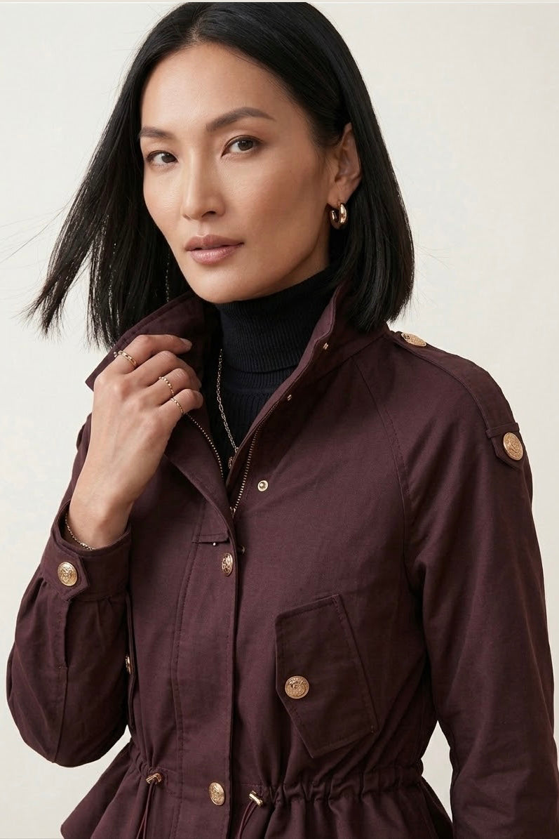 A woman wearing a dark plum-burgundy utility jacket with gold-tone buttons over a black turtleneck, photographed against a light neutral studio background.