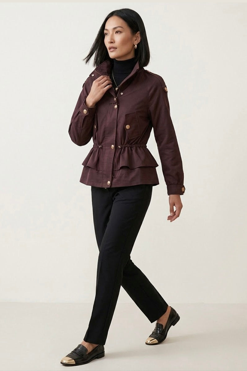 A woman modeling a dark plum-brown peplum jacket with gold buttons, worn over a black turtleneck and black pants, against a plain off-white studio background.