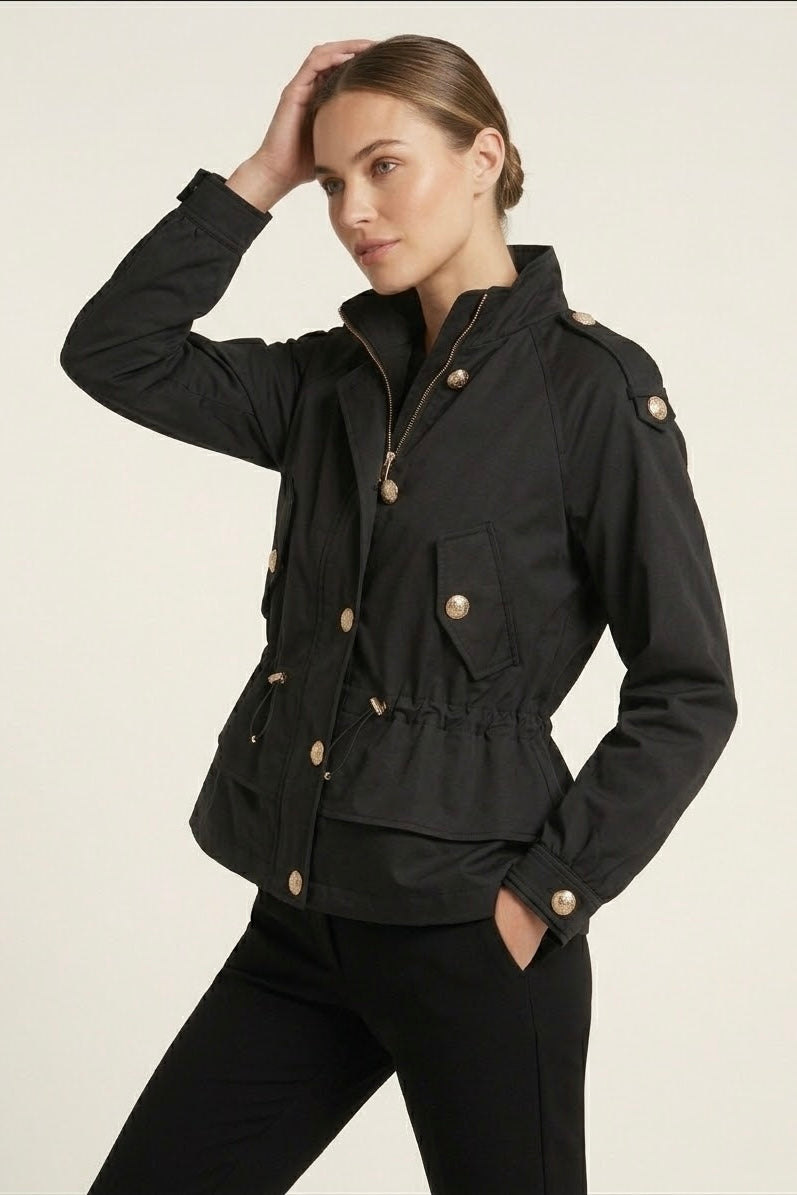 A woman modeling a black cropped utility jacket with gold-tone buttons and a high collar, paired with black pants, against a plain light background.