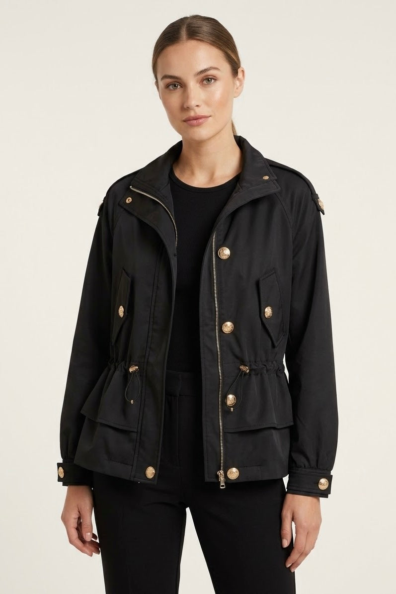 A woman modeling a black utility jacket with gold-tone buttons and zippers, worn over a black top and black pants, against a plain light beige studio background.