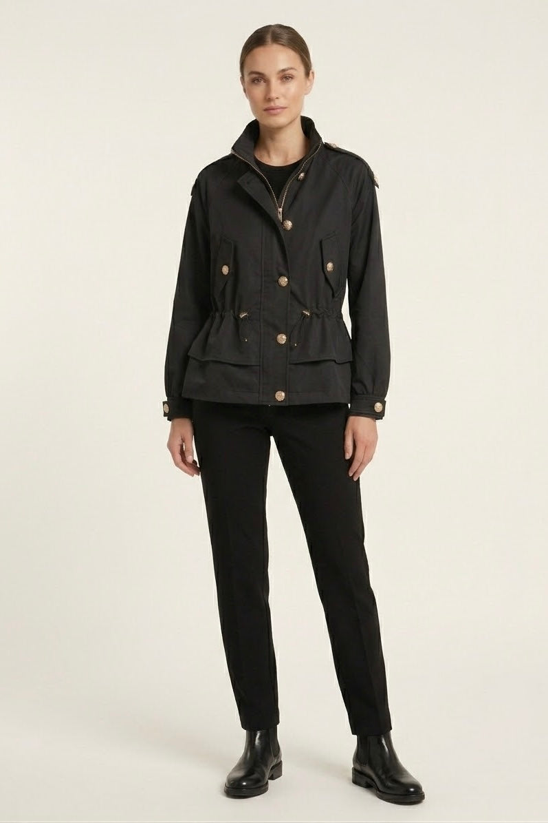 A model stands against a plain light background wearing a black cropped jacket with gold-tone buttons, black trousers, and black ankle boots.
